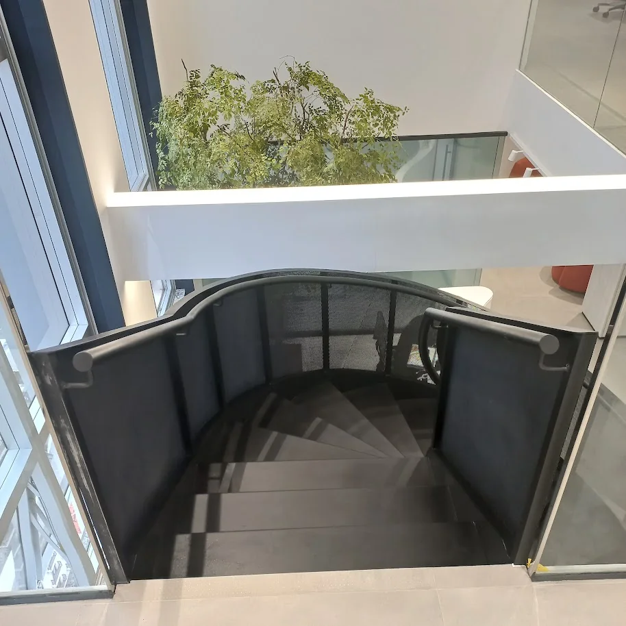 Vista aérea de escada helicoidal metálica preta com guarda-corpo de vidro e jardim interno ao fundo